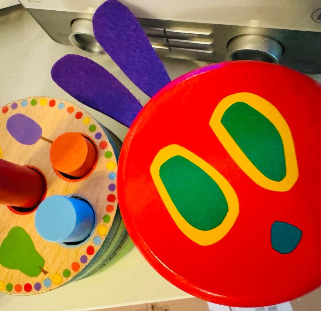 Holzspielzeug mit einem roten Deckel als Kopf der kleinen Raupe Nimmersatt und lila Filzohren neben einem bunten Holzsteckspiel.