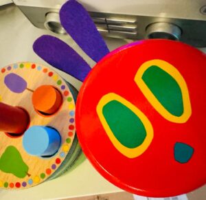 Holzspielzeug mit einem roten Deckel als Kopf der kleinen Raupe Nimmersatt und lila Filzohren neben einem bunten Holzsteckspiel.